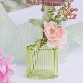 Grüne Mini-Glasvase mit floraler Dekoration auf weißem Tisch und weichem Stoff im Hintergrund.