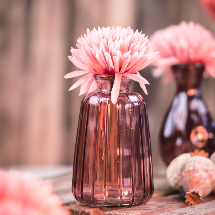 Glasvase Verone 11 cm Vintage Tischvase Rosa Grün Transparent Deko Geburtstag Tisch-Deko Hochzeit Geburtstag Vase aus Glas festliche Tischdeko Wohnaccessoire Glasvase dekorativ Rosa Glasvase Verone mit rosafarbener Blüte auf rustikalem Holztisch, im Hintergrund weitere Vasen in weicher Unschärfe.