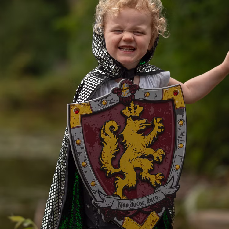 Ritterschild für Kinder EVA Schaumstoff mit Löwenwappen weich Mittelalter Kostüm-Zubehör Schutzschild Fasching Karneval Mottoparty Rollenspiel Kinderfasching Spielzeug-Schild Kind im Ritterkostüm hält einen Schaumstoff-Schild mit Löwenwappen vor dem Körper beim Spielen im Freien.