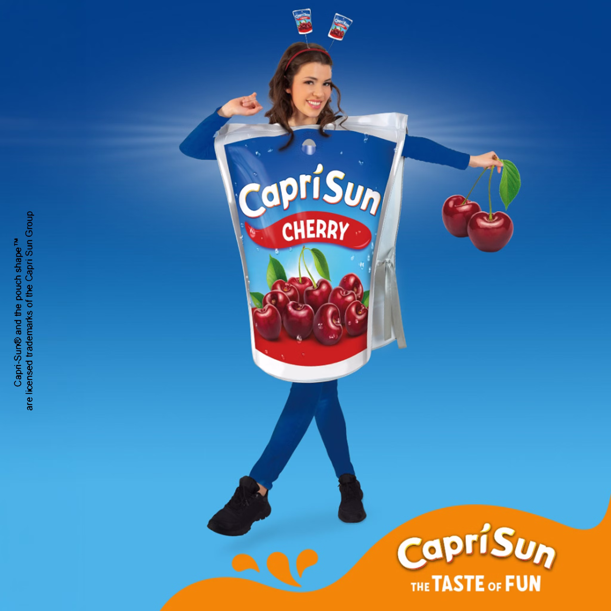 Frau im Capri Sun Cherry Kostüm mit passendem Haarreif und Kirsch-Dekoration vor blauem Capri Sun Hintergrund.