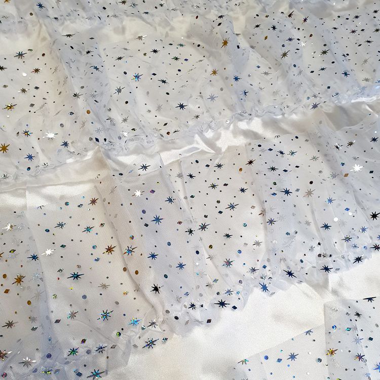 Herrenmodel trägt ein weißes Engel-Kleid aus Satin mit mehrlagigem Glitter-Tüll, kombiniert mit Engelsflügeln und Heiligenschein, vor weißem Hintergrund.